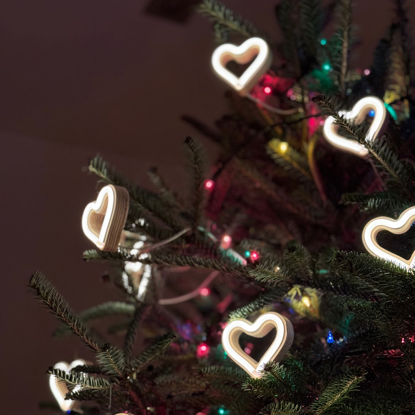 Multiple warm white LED neon heart ornaments decorating a Christmas tree with colorful lights - handmade neon heart holiday decor by Our Glowing Hearts
