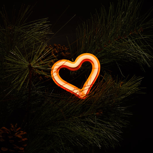 Orange neon heart ornament glowing on a Christmas tree with pine branches and pine cones - LED neon holiday decor by Our Glowing Hearts