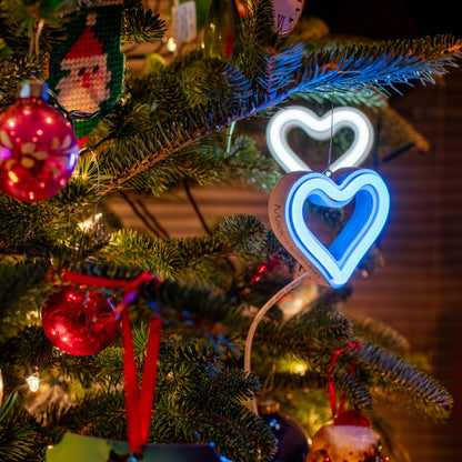 Cool white and cyan LED neon heart ornaments hanging on a decorated Christmas tree with festive ornaments and lights by Our Glowing Hearts