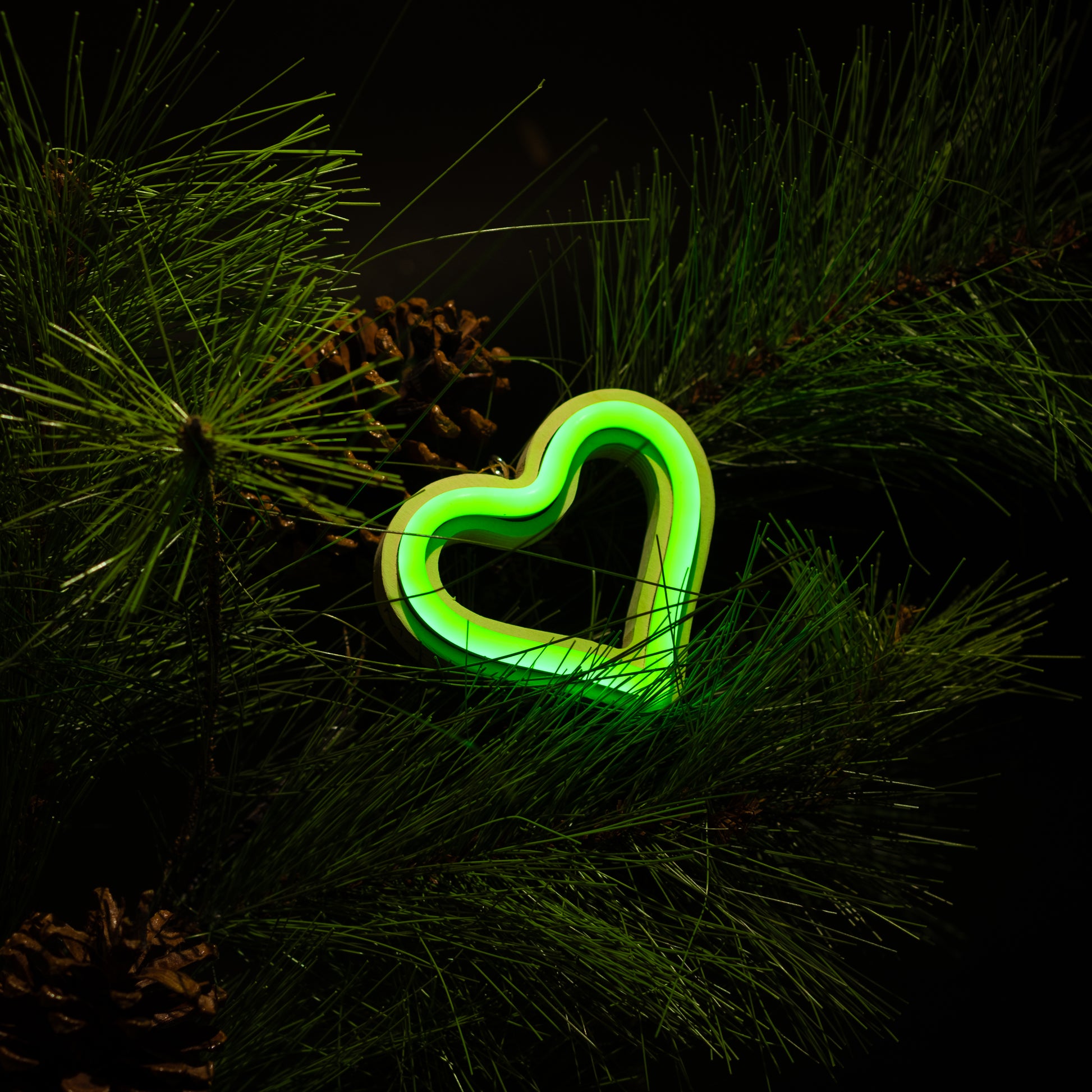Green neon heart ornament hanging on a Christmas tree with pine branches and pine cones - LED neon holiday decor by Our Glowing Hearts