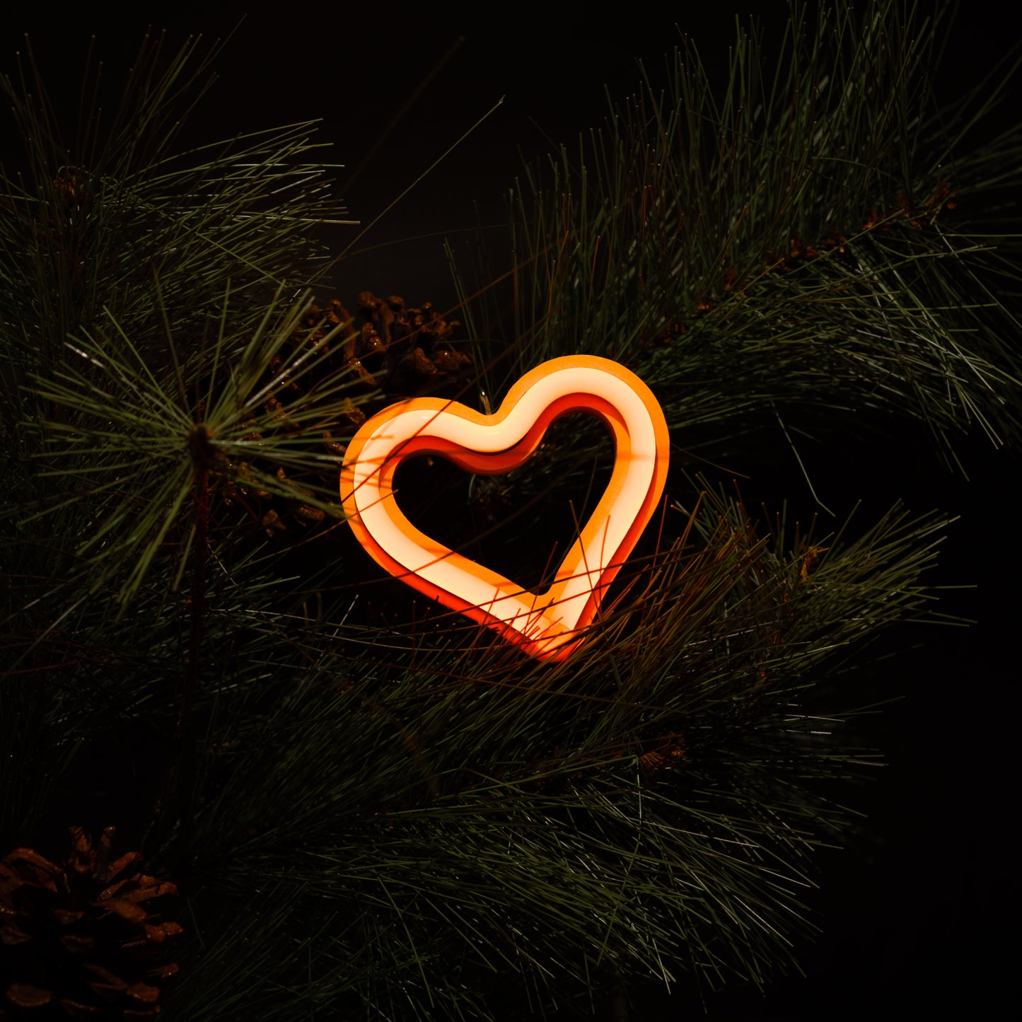 Orange neon heart ornament glowing on a Christmas tree with pine branches and pine cones - LED neon holiday decor by Our Glowing Hearts