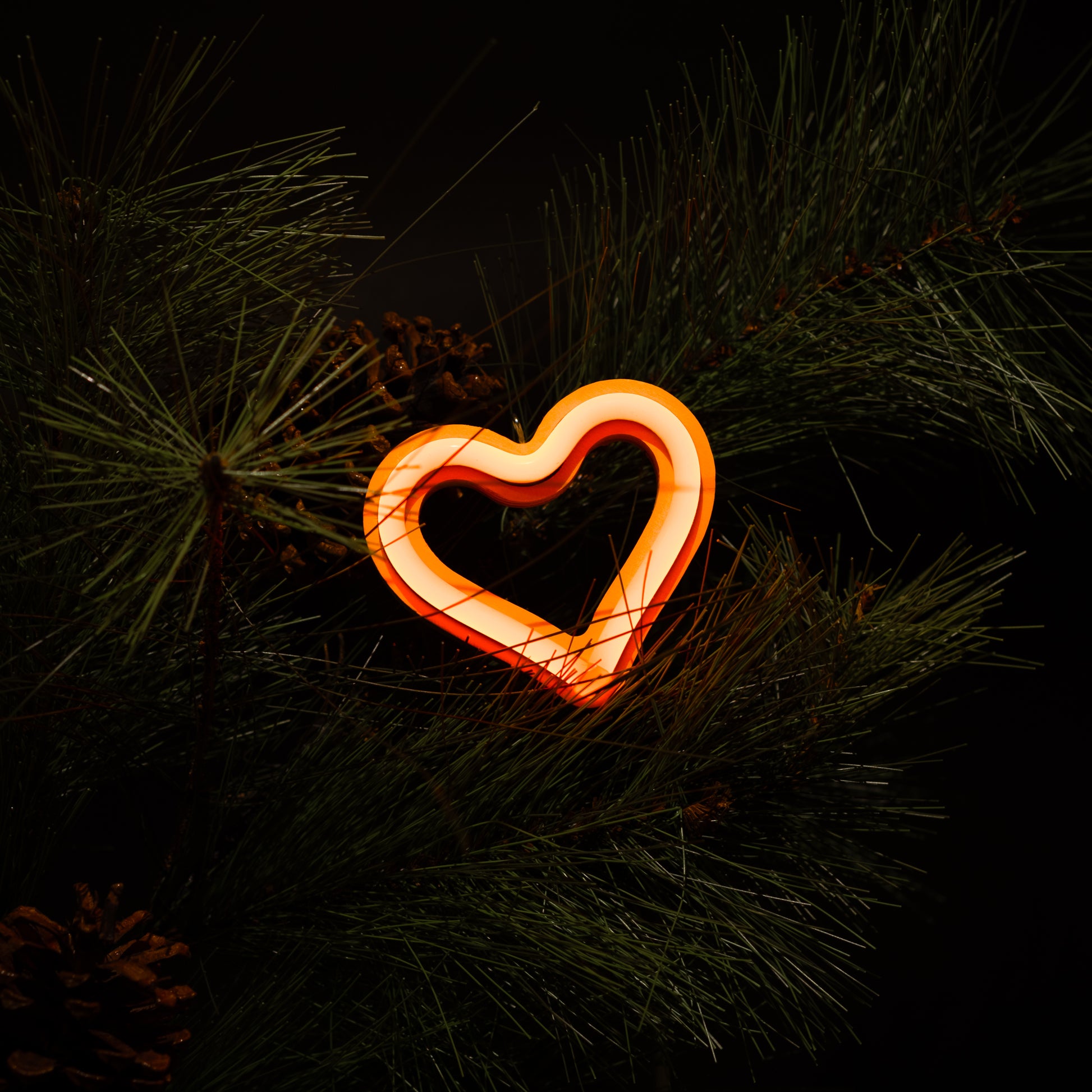 Orange neon heart ornament glowing on a Christmas tree with pine branches and pine cones - LED neon holiday decor by Our Glowing Hearts