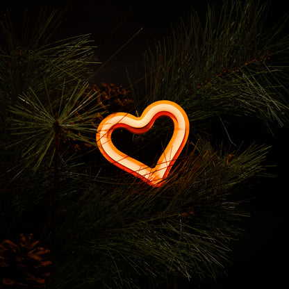 Orange neon heart ornament glowing on a Christmas tree with pine branches and pine cones - LED neon holiday decor by Our Glowing Hearts
