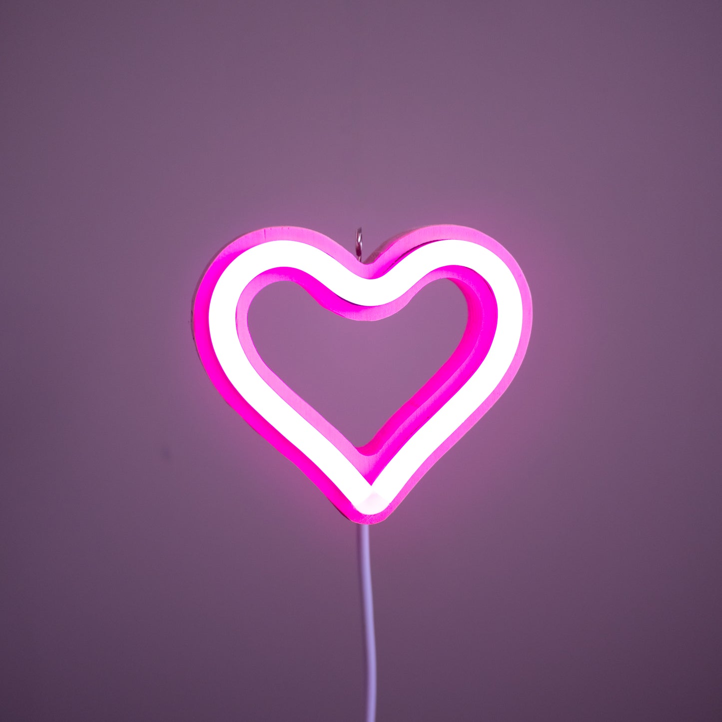 Front view of pink neon heart ornament with hanging hook glowing against a pink background - 3 inch LED neon heart light by Our Glowing Hearts