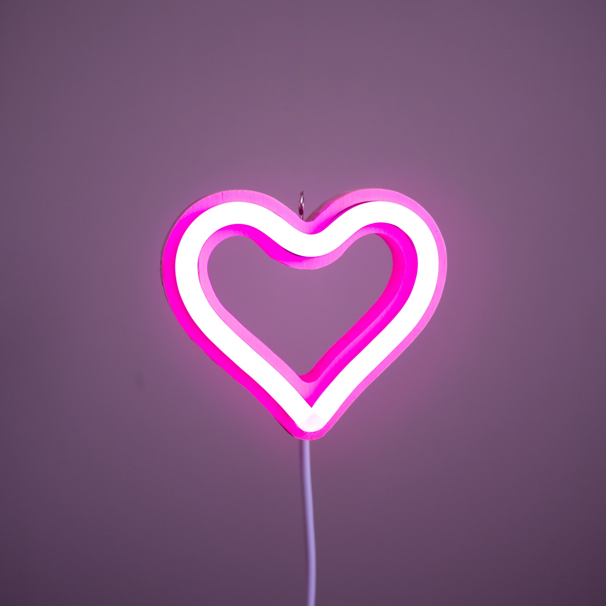 Front view of pink neon heart ornament with hanging hook glowing against a pink background - 3 inch LED neon heart light by Our Glowing Hearts