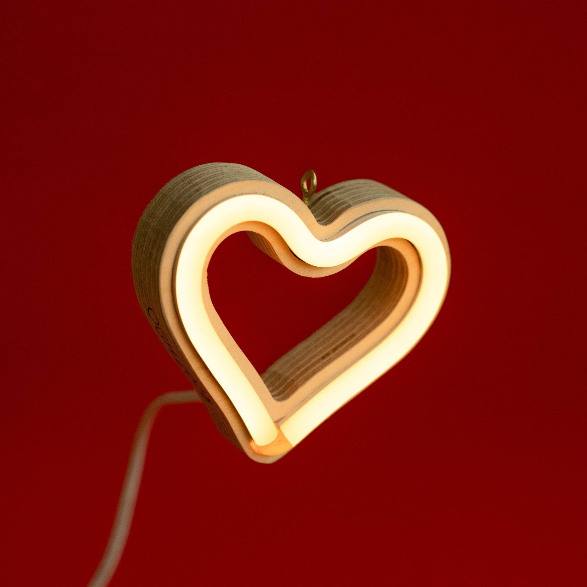 Front view of warm white neon heart ornament with hanging hook glowing against a red background - 3 inch LED neon heart light by Our Glowing Hearts