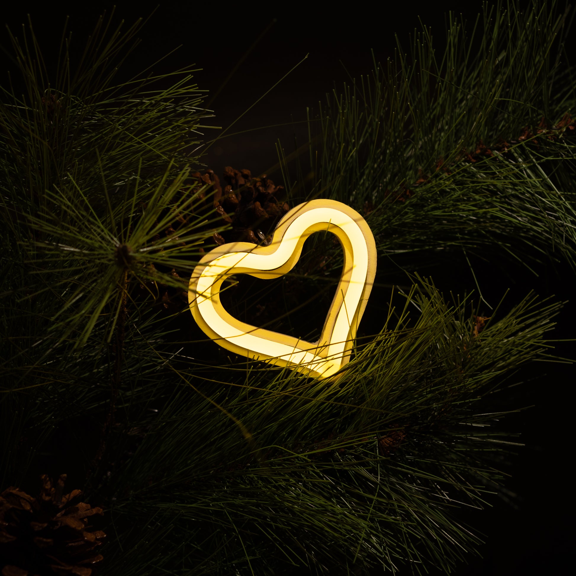 Yellow neon heart ornament hanging on a Christmas tree with pine branches and pine cones - LED neon holiday decor by Our Glowing Hearts