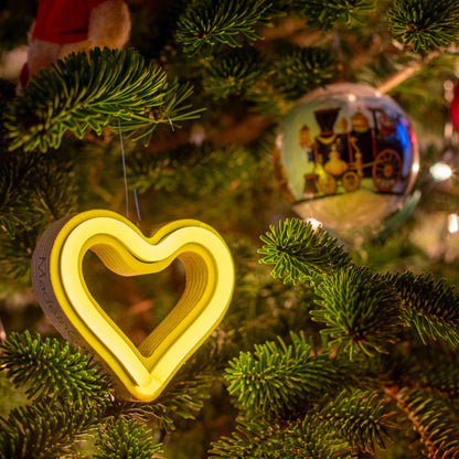 Close-up of yellow neon heart ornament glowing on a Christmas tree with glass bauble and pine branches - LED neon holiday decor by Our Glowing Hearts
