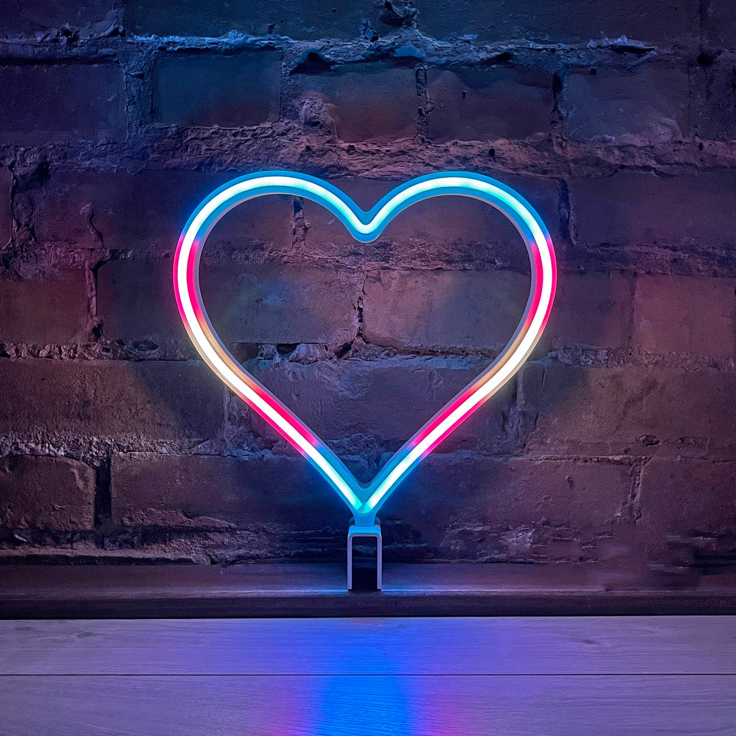 Neon Queer Heart art in transgender pride flag colors glowing against a brick wall - 12 inch LED neon heart light with 12 pride flag modes by Our Glowing Hearts, handmade in Canada