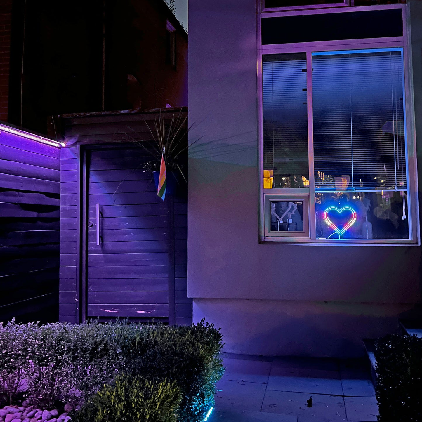 Neon Queer Heart art displayed in a home window at night with pride flag on the house - LED neon heart window display by Our Glowing Hearts