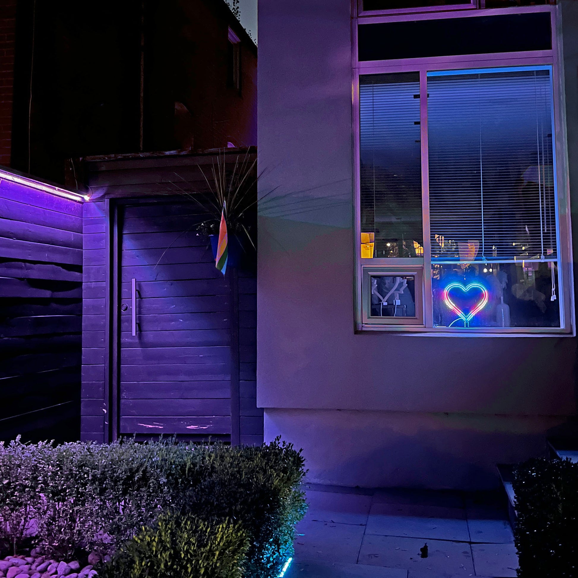 Neon Queer Heart art displayed in a home window at night with pride flag on the house - LED neon heart window display by Our Glowing Hearts