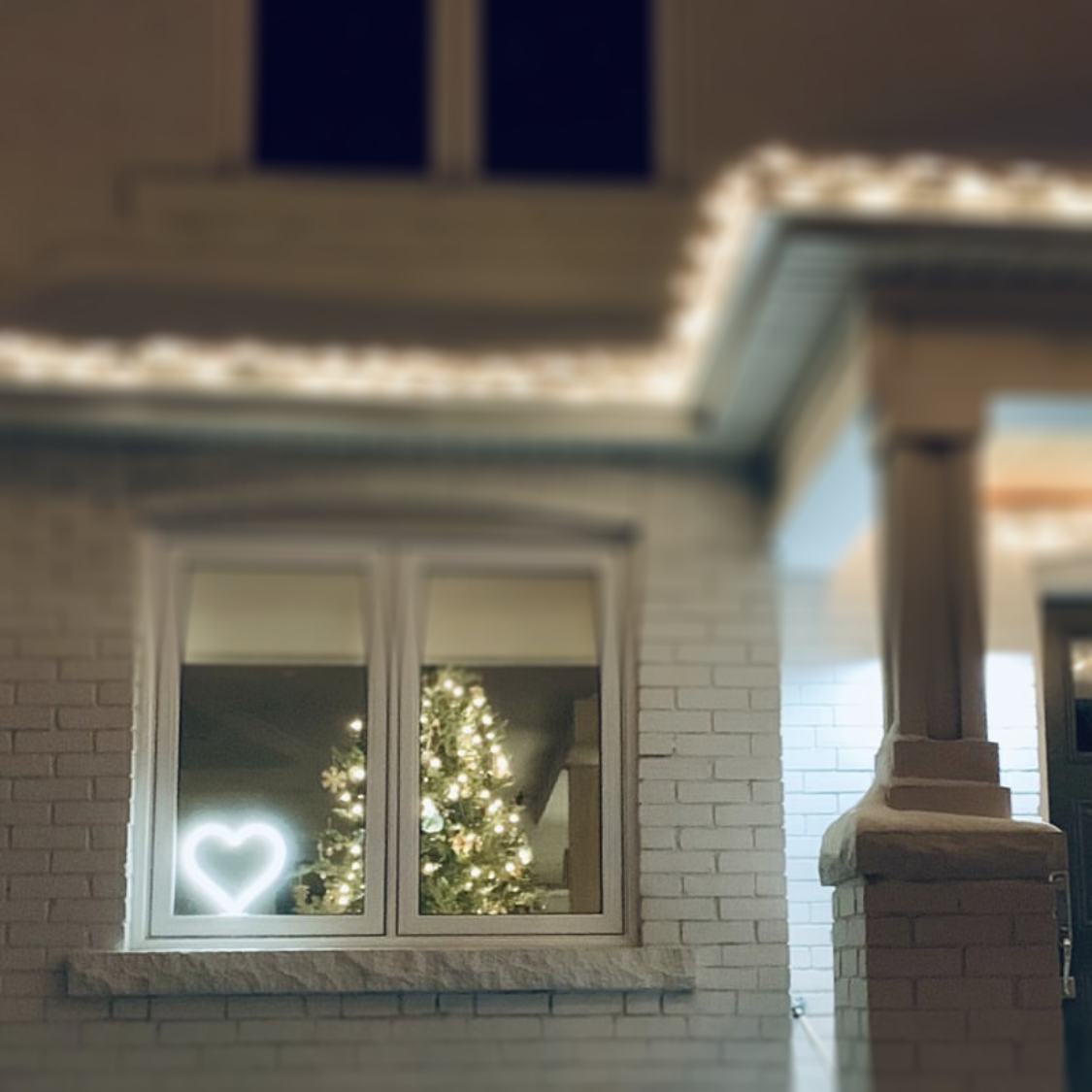 Cool white neon heart art displayed in a window next to a Christmas tree at night - LED neon heart holiday window decor on a brick house exterior by Our Glowing Hearts