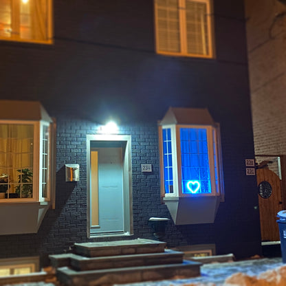 Cyan neon heart art glowing in a bay window of a brick house at night - LED neon heart visible from the street, perfect window decor by Our Glowing Hearts