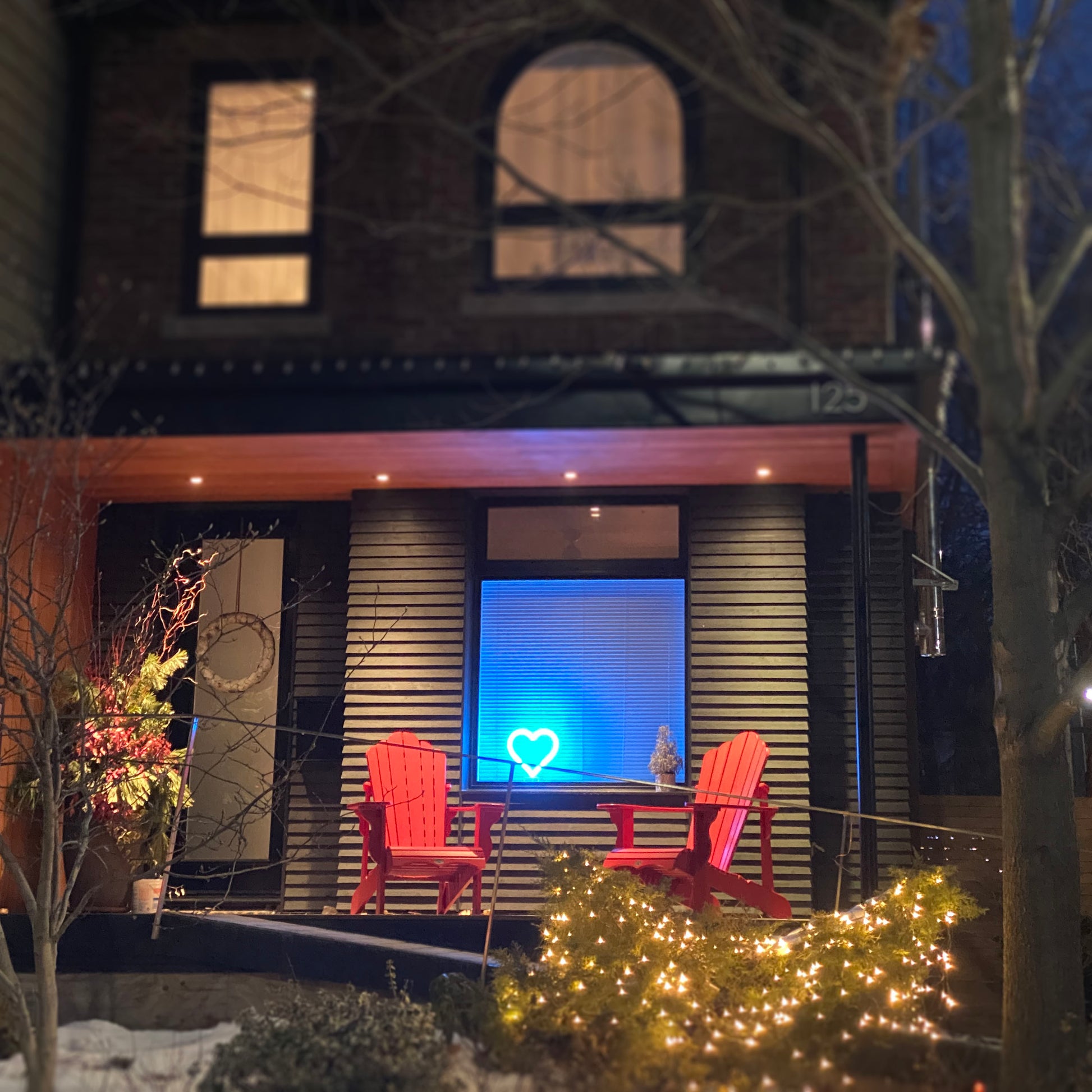 Cyan neon heart art glowing in a home window at night with cozy porch and string lights - LED heart-shaped light spreading love in the neighborhood by Our Glowing Hearts