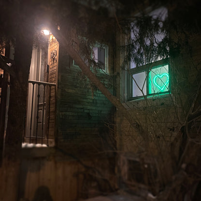 Green neon heart art glowing in a home window at night - LED heart-shaped light visible from outside, spreading love in the neighborhood by Our Glowing Hearts