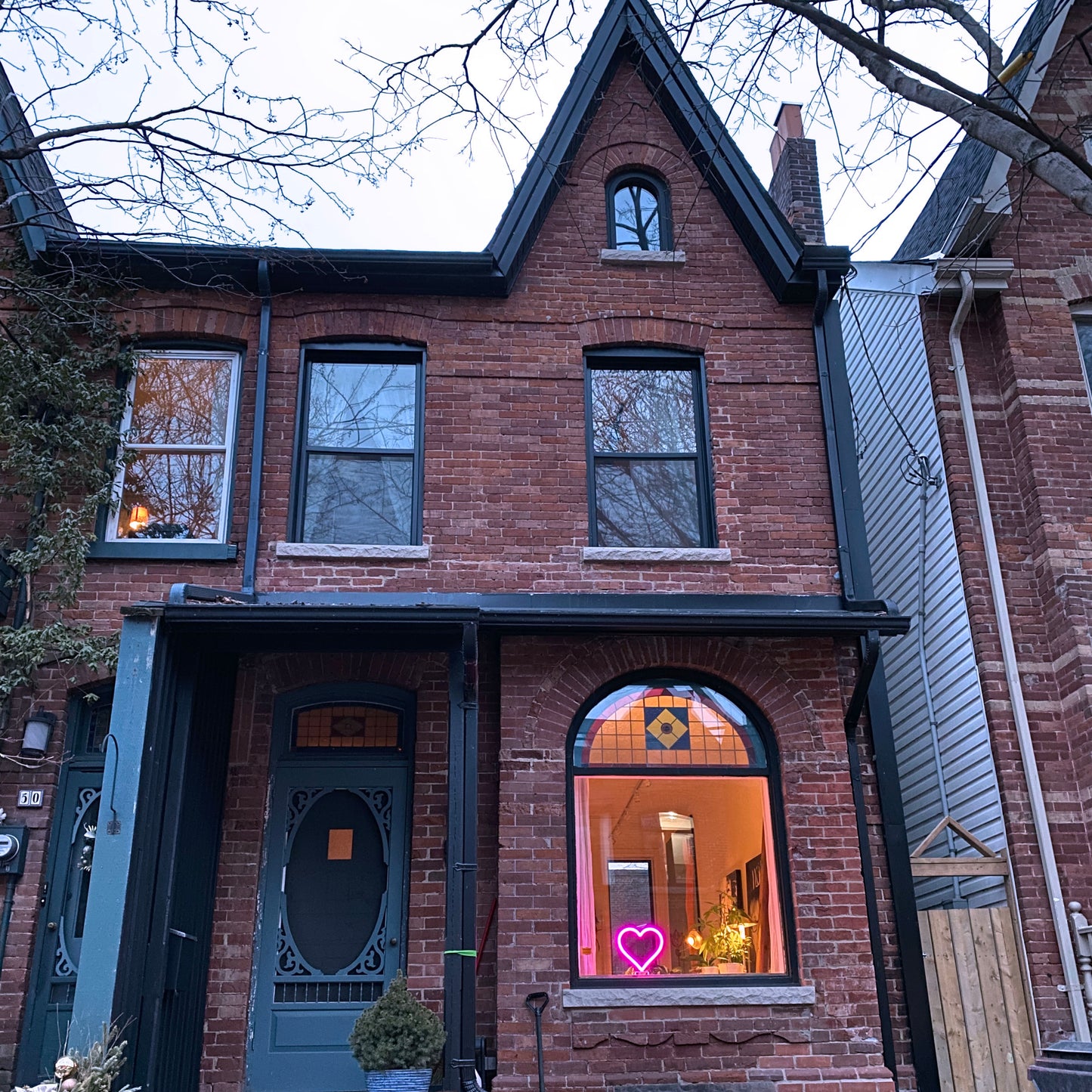 Magenta neon heart art glowing in the front window of a brick home at dusk - handmade LED heart light spreading love in the neighborhood by Our Glowing Hearts