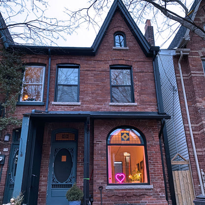Magenta neon heart art glowing in the front window of a brick home at dusk - handmade LED heart light spreading love in the neighborhood by Our Glowing Hearts